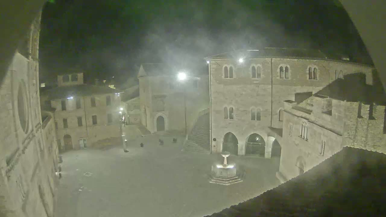 A nighttime view of a town square with ancient stone buildings surrounding a central fountain, under foggy or hazy conditions.