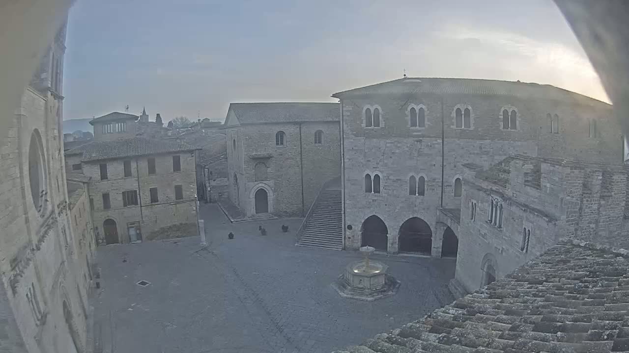 A high-angle, hazy view shows a town square with several stone buildings surrounding a fountain, under a pale, overcast sky.