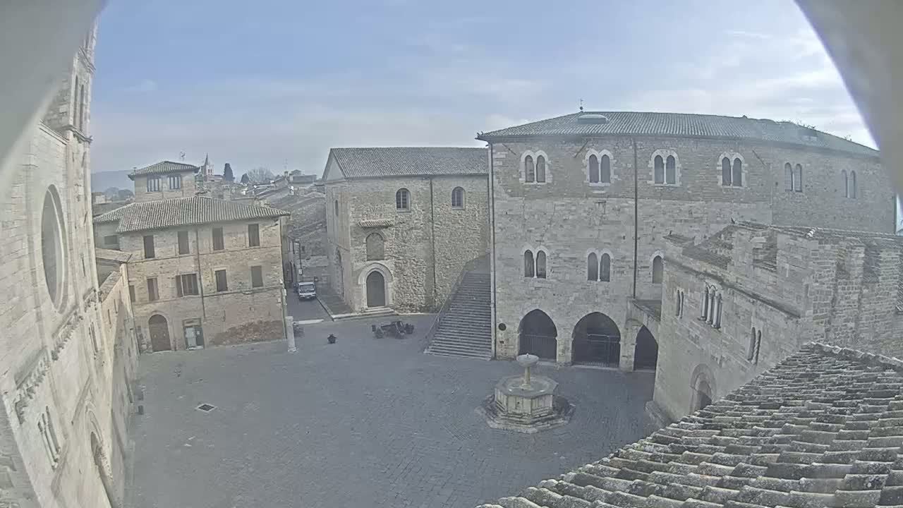A stone plaza, under a mostly clear sky, is surrounded by medieval-style buildings, one featuring a fountain in its center.