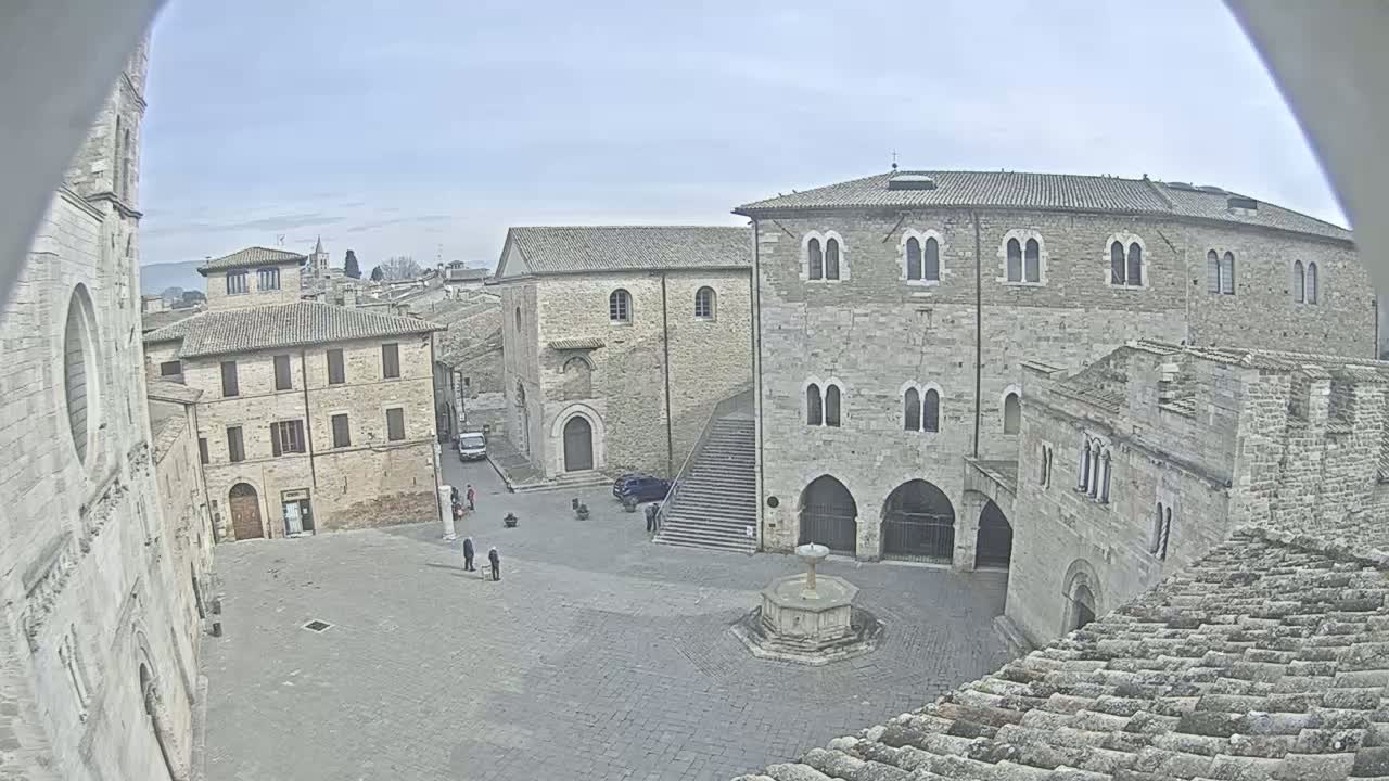 A mostly empty stone plaza, under a cloudy sky, is surrounded by medieval-style buildings, with one featuring a prominent staircase and a central fountain.