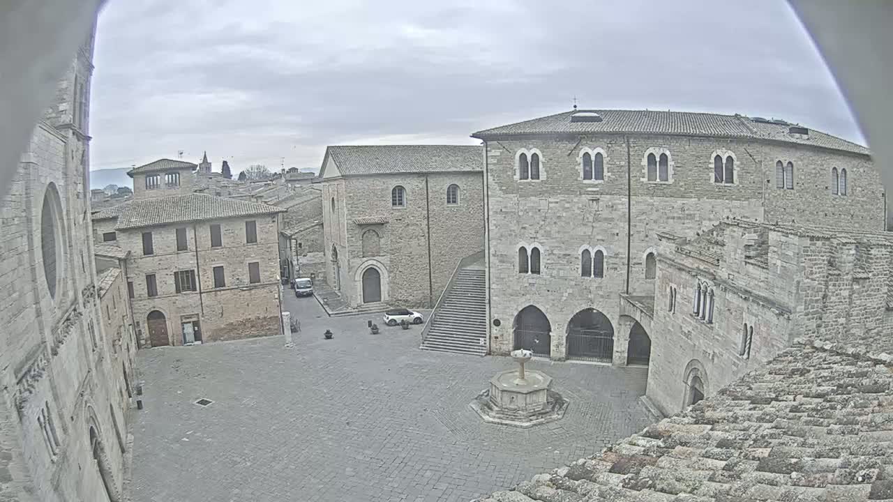 A mostly empty stone plaza in a historic town on an overcast day, surrounded by medieval buildings, one featuring a fountain at its center.