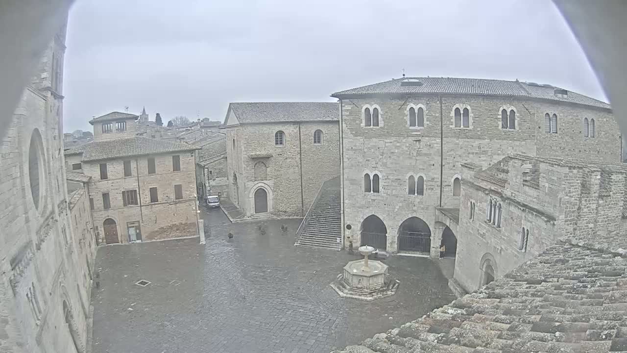 A wet stone plaza, surrounded by medieval-style buildings under an overcast sky, features a central fountain.