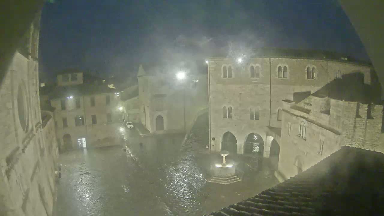 A town square, flooded with rainwater under a dark, stormy night sky, is surrounded by old stone buildings.