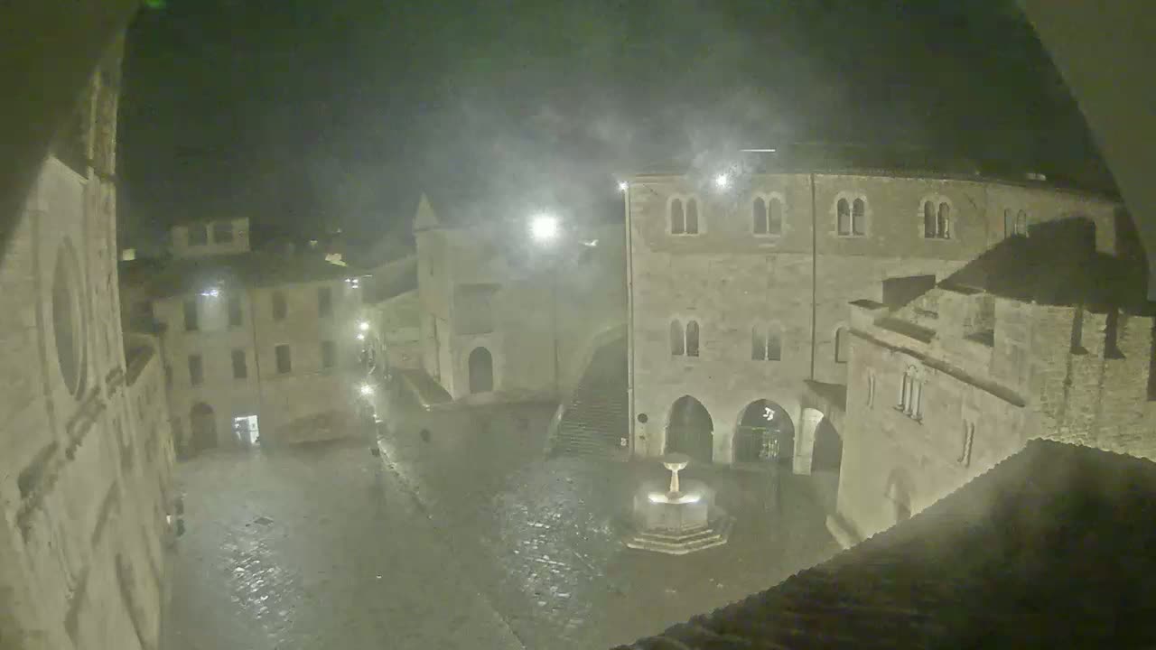 A nighttime view of a rain-swept town square, featuring ancient stone buildings and a fountain at its center, under a hazy sky.