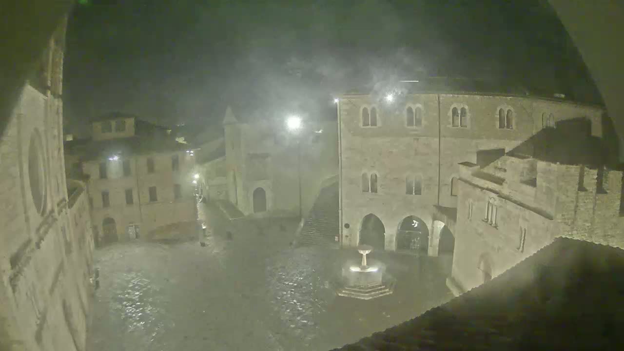 A nighttime view of a town square with old stone buildings, a fountain in the center, and misty, rainy weather.