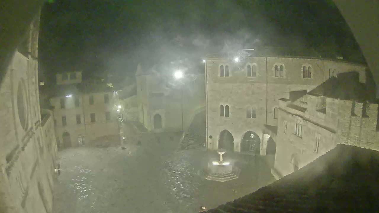 A nighttime view of a town square with ancient stone buildings, a central fountain, and a misty or foggy atmosphere.