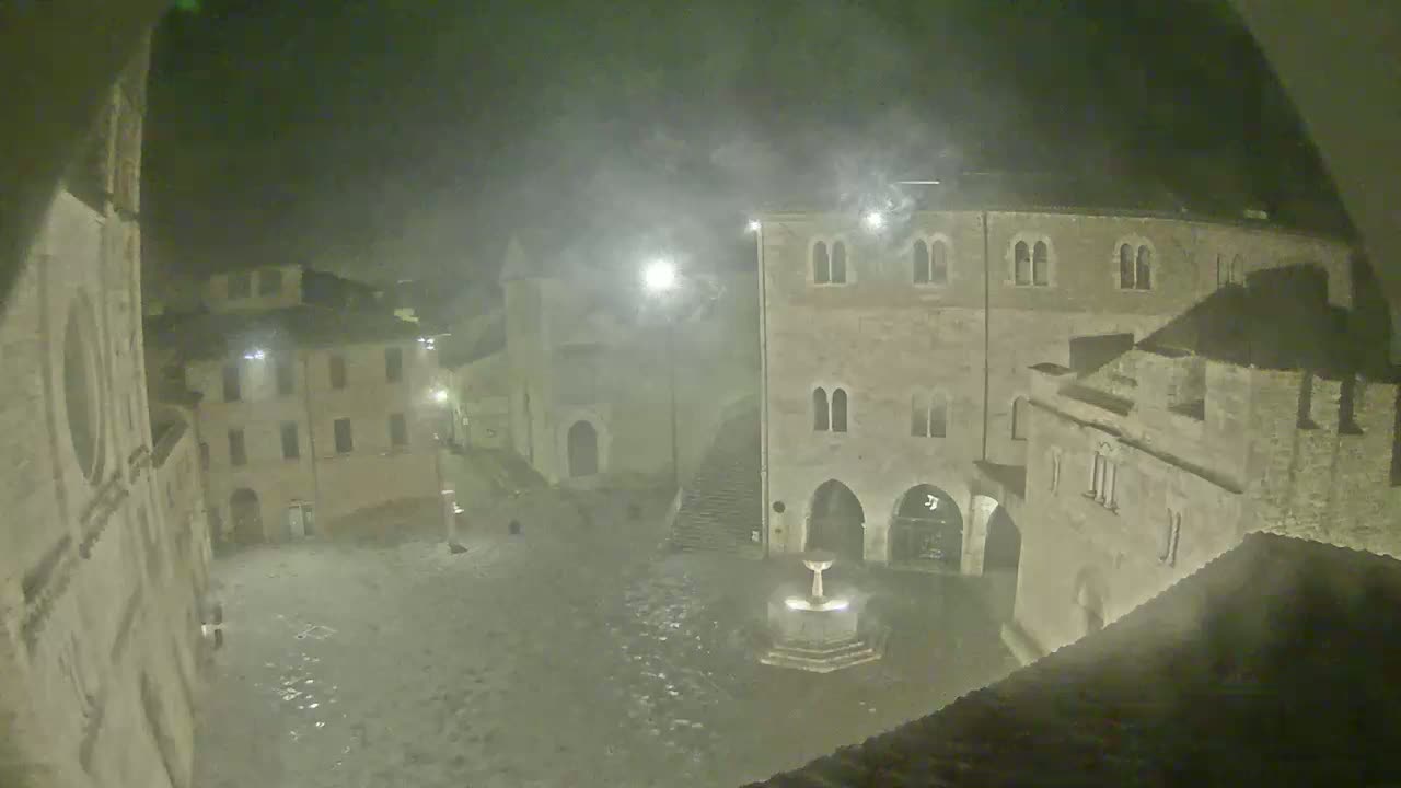 A nighttime view of a town square with stone buildings and a fountain, under hazy or foggy conditions.