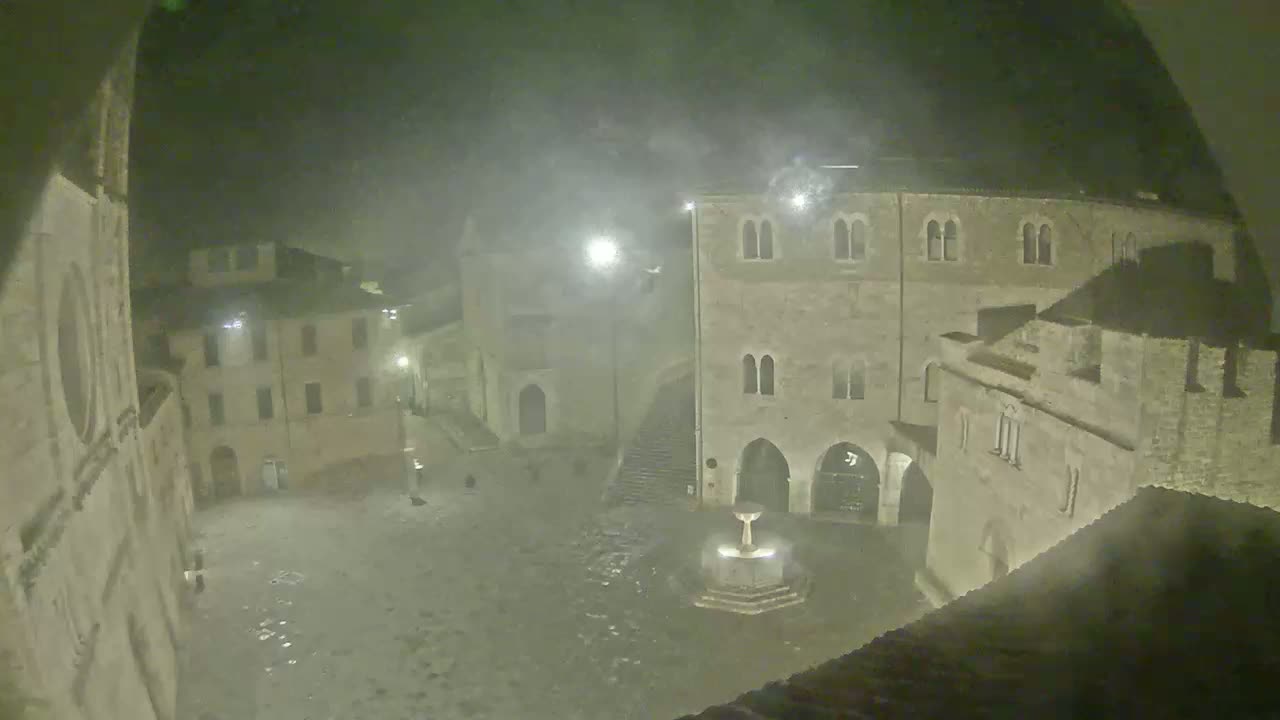 A nighttime view of a town square with old stone buildings and a fountain, under hazy or foggy conditions.