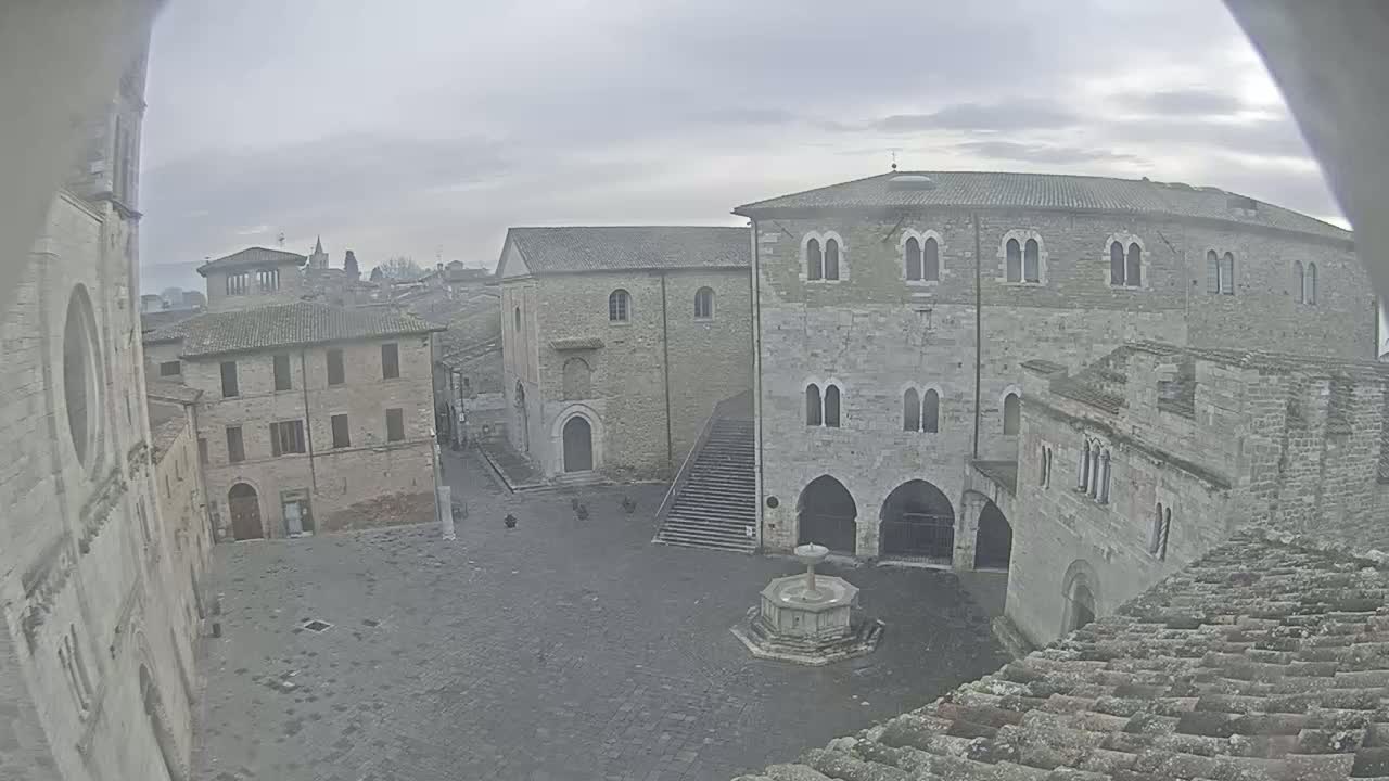 A stone plaza with a fountain is surrounded by medieval-style buildings on an overcast day.