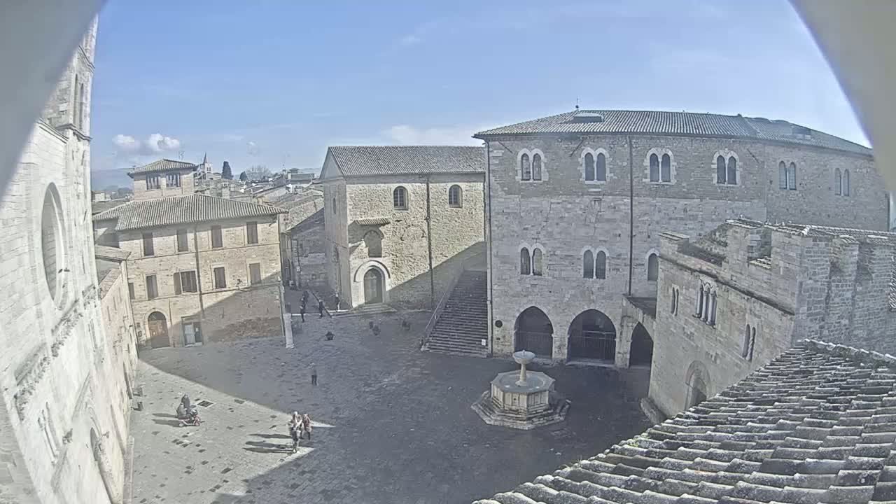 A sunny day reveals a high-angle view of a town square surrounded by old stone buildings, one with a central fountain.