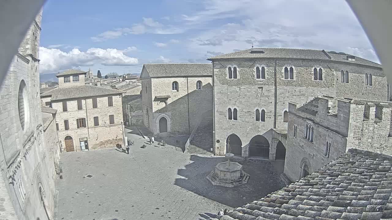 A mostly empty town square with several stone buildings, a fountain, and a stairway under a partly cloudy sky.