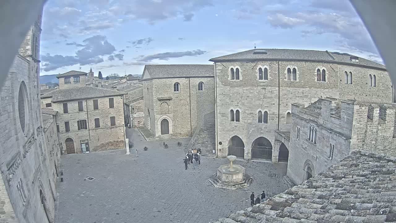 A high-angle view shows a town square with several stone buildings, a fountain, and a small group of people under a partly cloudy sky.