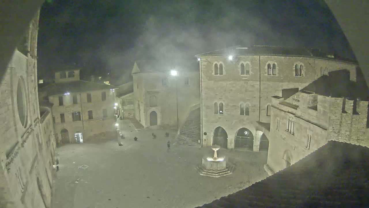 A nighttime view of a town square with stone buildings and a fountain, under hazy or foggy conditions.