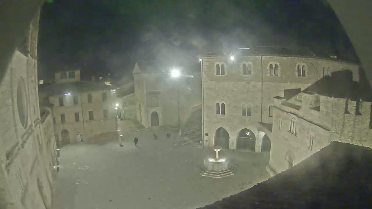A nighttime view of a town square with several stone buildings surrounding a fountain, under hazy or foggy conditions.