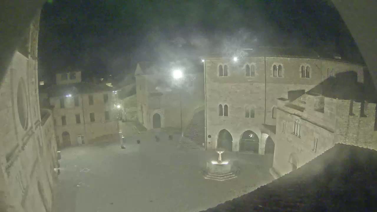 A nighttime view of a town square with stone buildings, a fountain, and hazy atmospheric conditions.