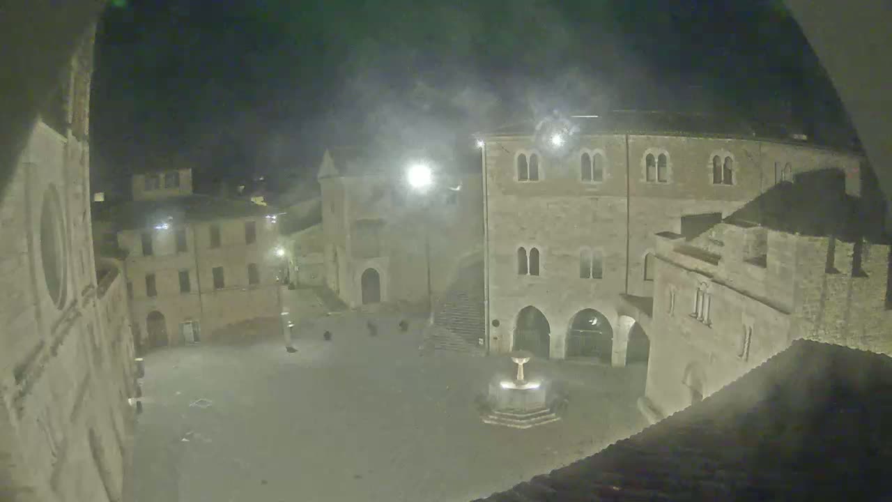 A nighttime, hazy view of a town square, centered around a fountain, surrounded by aged stone buildings.