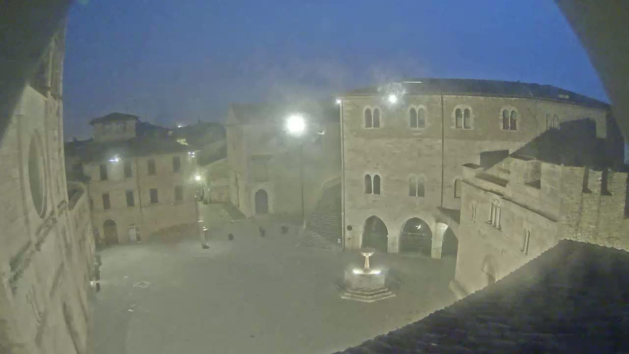 A nighttime view of a town square with ancient stone buildings under hazy, dark conditions.