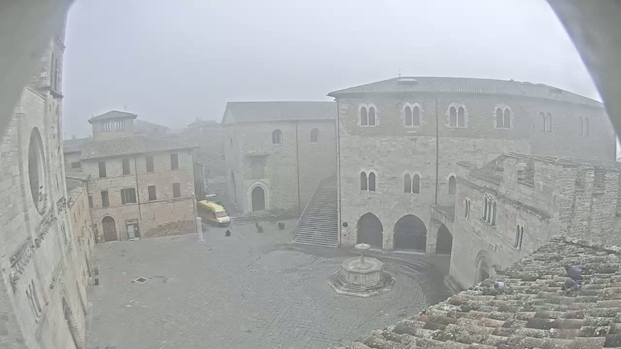 A foggy view of a town square, surrounded by ancient stone buildings, with a fountain in the center and a vehicle parked nearby.