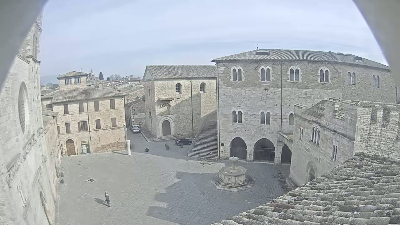 A mostly empty town square with a fountain in the center is surrounded by aged stone buildings under a bright, overcast sky.