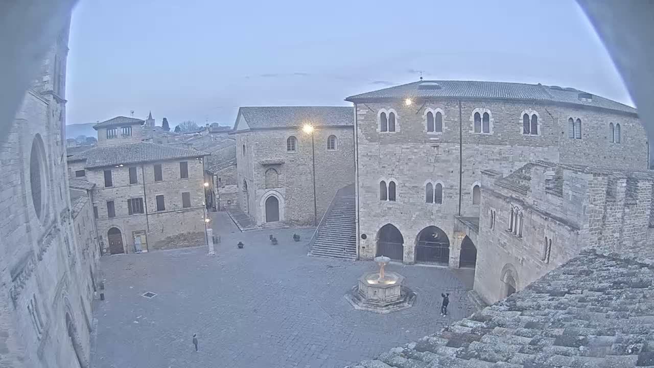 A stone plaza with a fountain is surrounded by medieval-style buildings under a twilight sky.