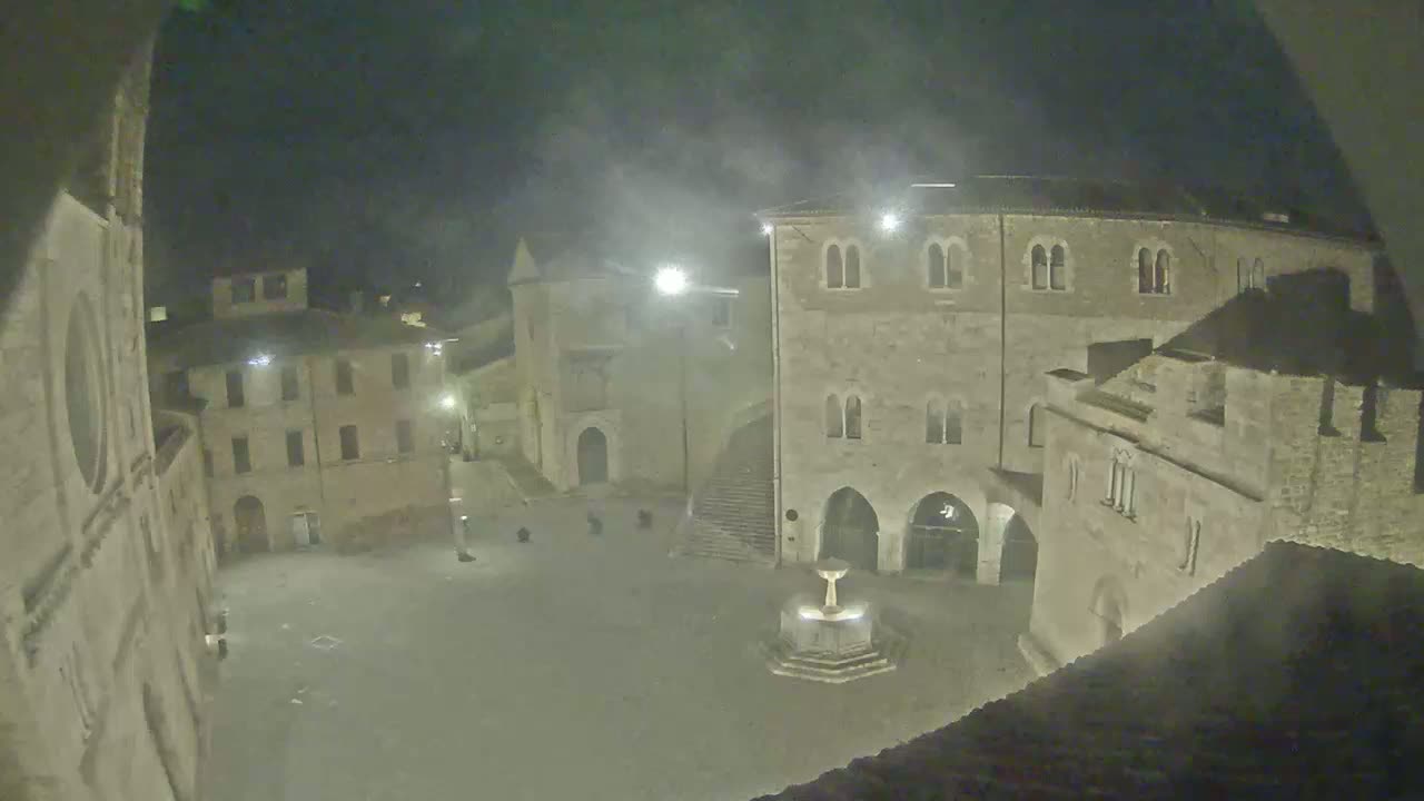 A nighttime view of a town square, shrouded in mist, features ancient stone buildings surrounding a central fountain.