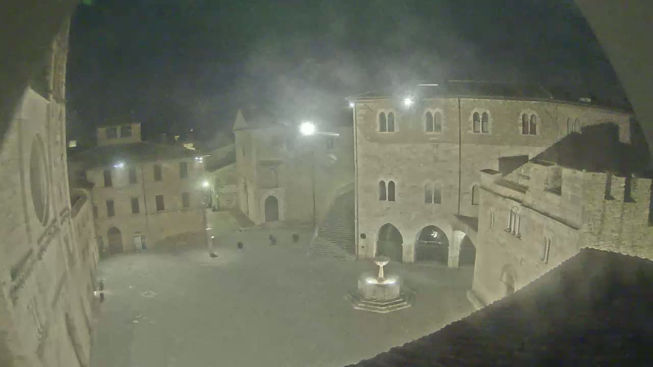 A nighttime view of a town square, possibly in Europe, with stone buildings surrounding a central fountain, under hazy or foggy conditions.
