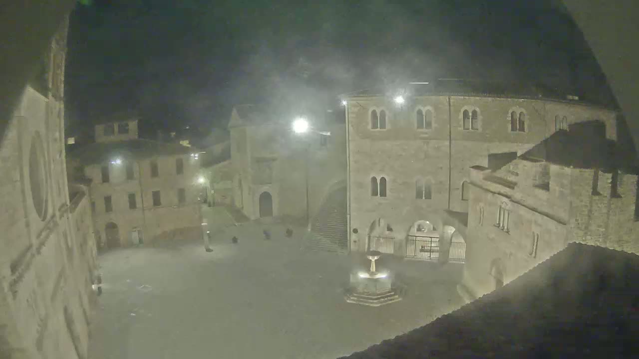A nighttime, possibly foggy, view of a town square surrounded by stone buildings and a fountain in the center.