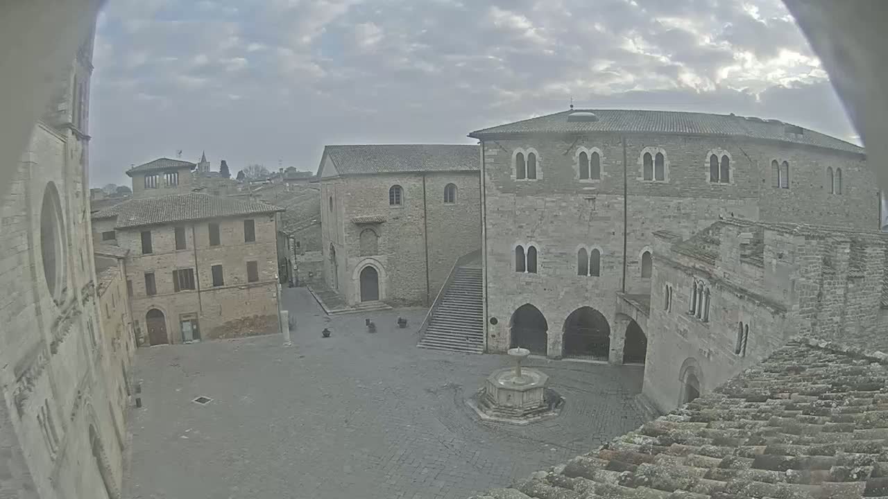 A mostly empty stone plaza with a fountain, surrounded by medieval-style buildings under an overcast sky.