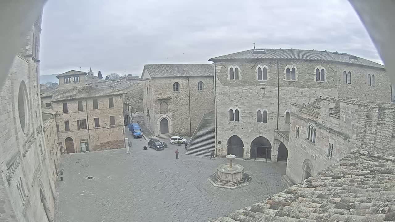 A stone plaza with a fountain in the center, surrounded by medieval-looking buildings, on an overcast day.