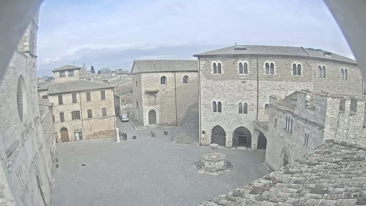 A stone plaza in a historic town on an overcast day, surrounded by aged buildings including a large stone structure with arched entryways and a smaller building with a simple facade.