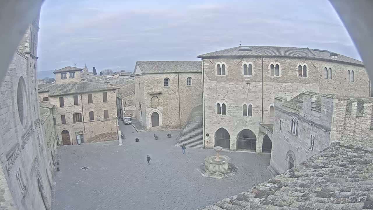 A high-angle view of a town square with several stone buildings, a fountain, and a few people, under an overcast sky.