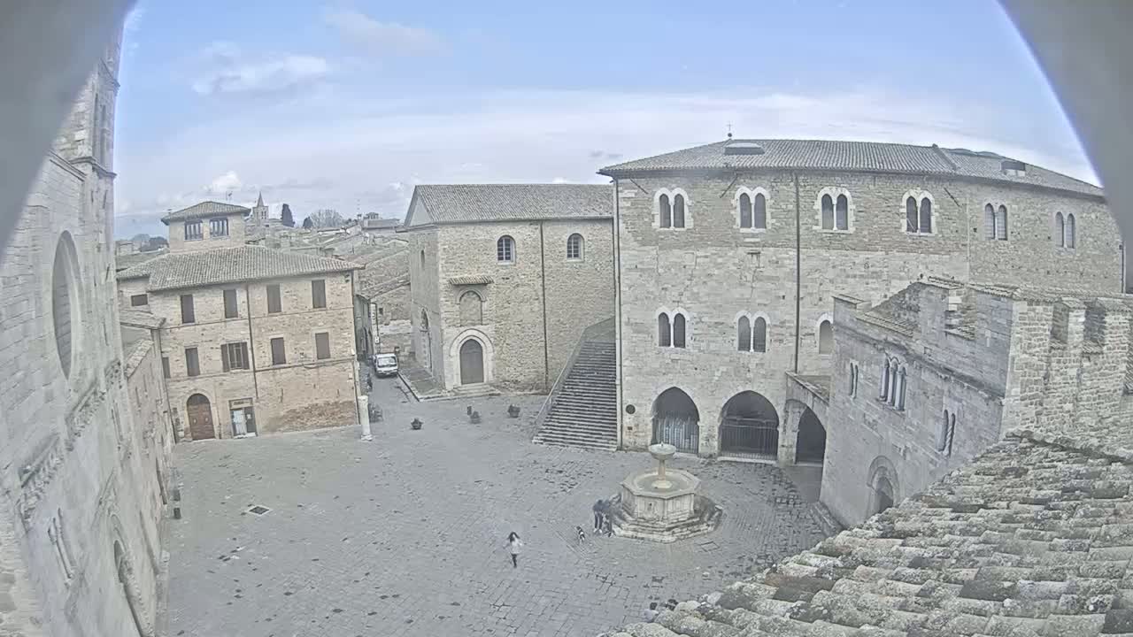 A high-angle view of a stone plaza in a historic town on a partly cloudy day, featuring several old buildings, a fountain, and a few people.