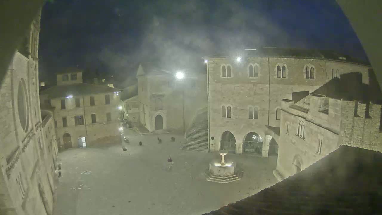 A nighttime, hazy view of a town square with stone buildings surrounding a fountain, a few people are sparsely scattered in the square.