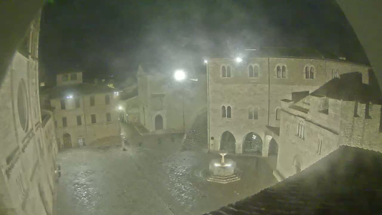 A nighttime, misty view of a cobblestone town square with a fountain, surrounded by old stone buildings.