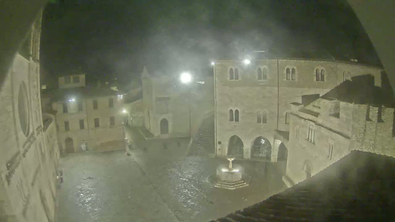 A nighttime view of a town square with old stone buildings, a fountain, and light rain or mist.