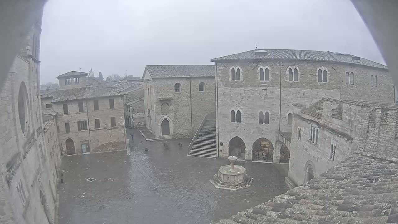 A rain-soaked town square is surrounded by medieval stone buildings, a fountain sits in the center, and a few people are visible.