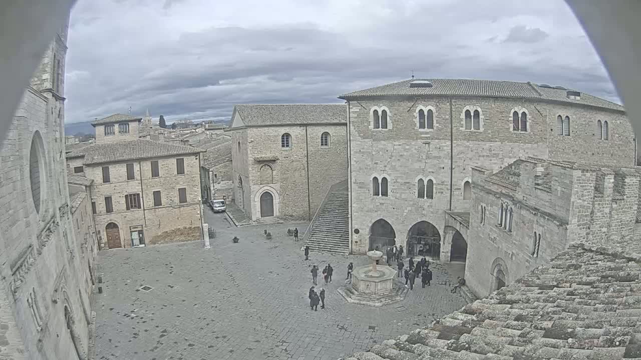 A high-angle view of a town square with several stone buildings, a fountain, and a few pedestrians under an overcast sky.