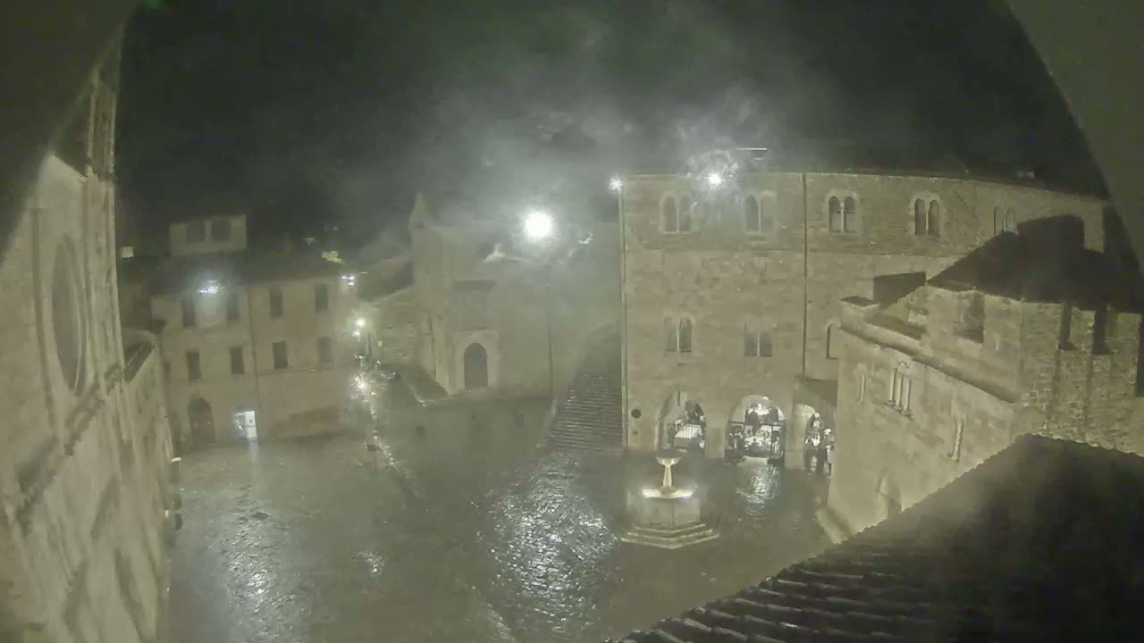 A floodlit town square, submerged in rainwater at night, is surrounded by ancient stone buildings.