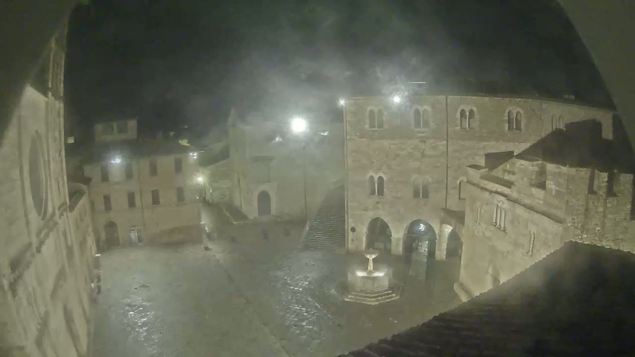 A nighttime view of a town square with ancient stone buildings, a fountain, and misty weather conditions.