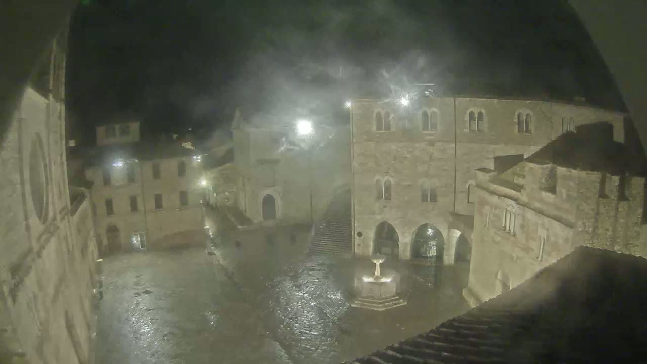A nighttime view of a town square, with stone buildings surrounding a fountain, during a heavy rain or windstorm.