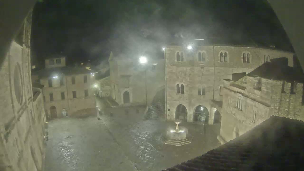 A nighttime view of a stone plaza with a fountain in a historic town, under misty or foggy conditions.