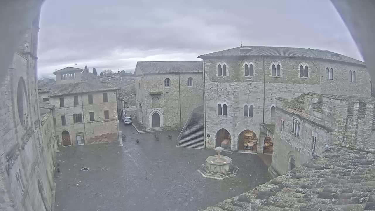 A stone plaza with a fountain is surrounded by medieval-style buildings under a cloudy sky.