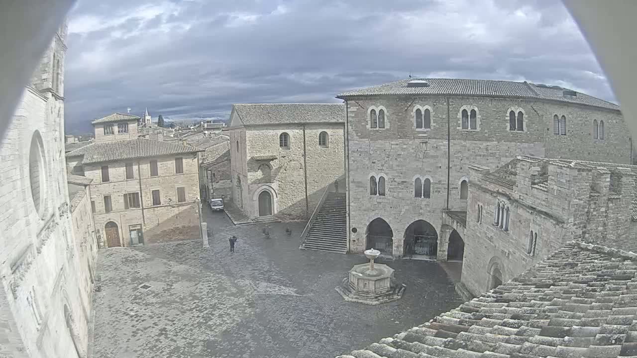 A mostly cloudy day reveals a stone plaza in a medieval-looking town, centered around a fountain, with several buildings surrounding it.