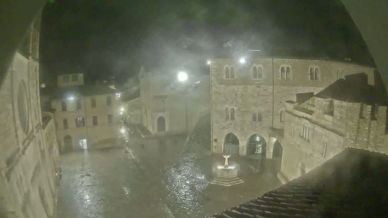 A town square, at night under heavy rain, is flooded around a central fountain, with old stone buildings surrounding it.