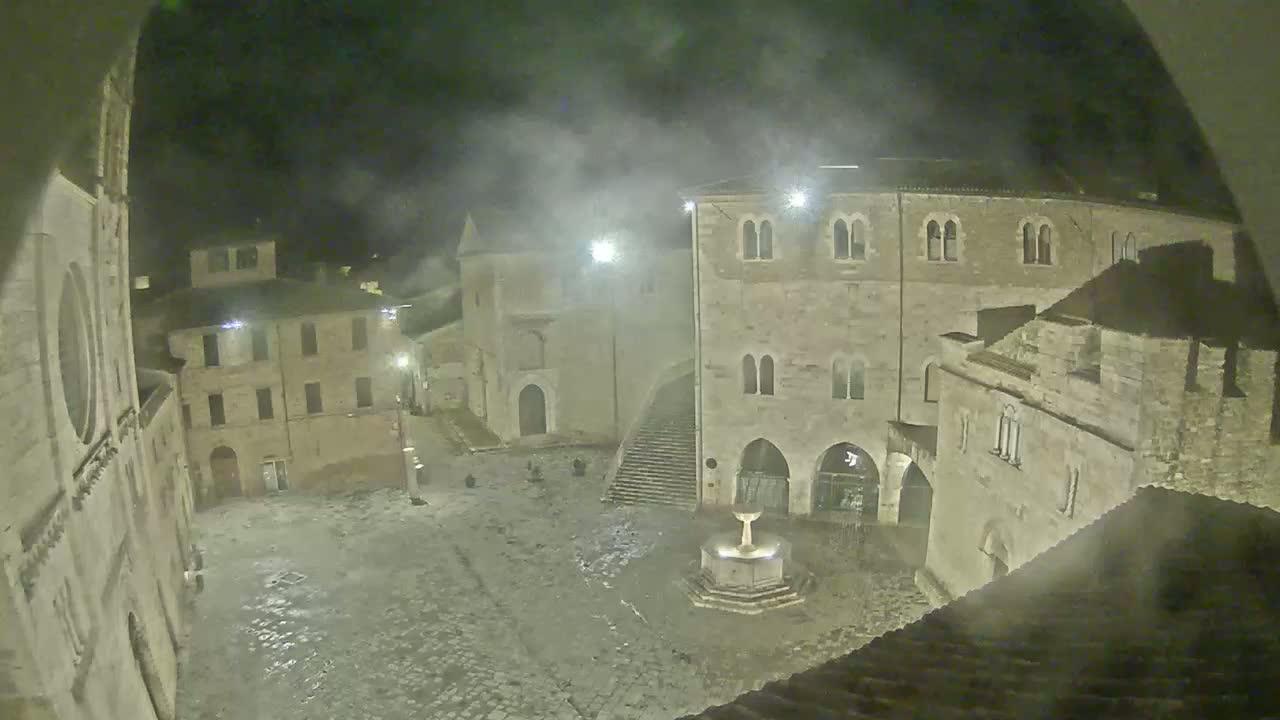 A nighttime view of a cobblestone town square with stone buildings surrounding a fountain, under hazy or foggy conditions.