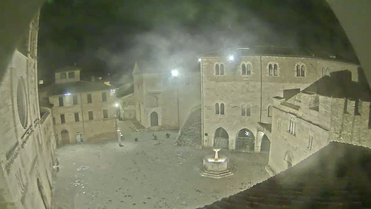 A nighttime, hazy view of a town square with several stone buildings surrounding a central fountain.