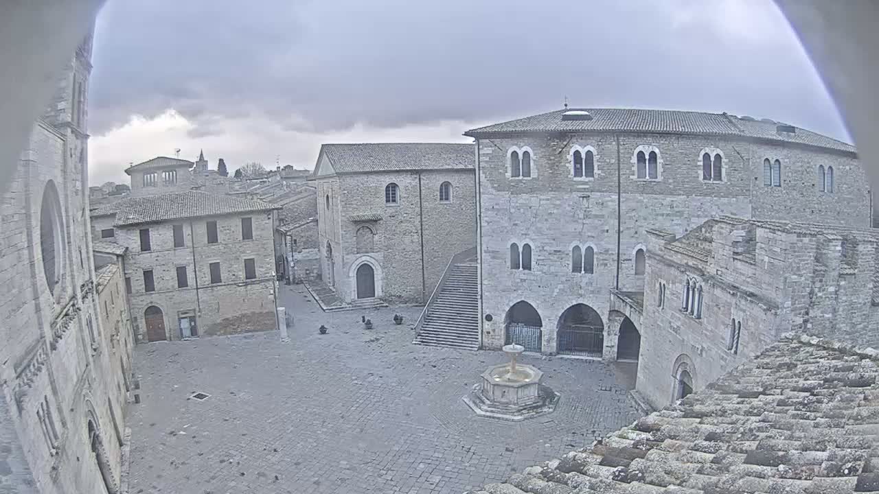 A stone plaza with a fountain, surrounded by medieval-style buildings under an overcast sky.
