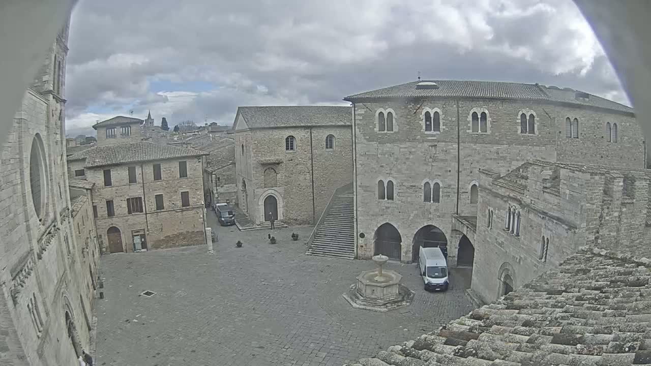 A stone plaza in a historic town, under a cloudy sky, is centered around a fountain with several buildings surrounding it, including a large stone structure with arched entryways.