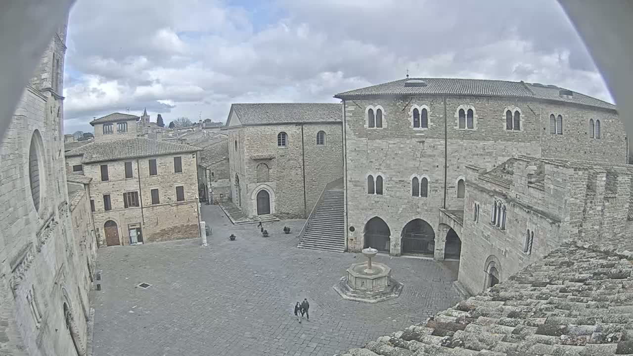 A stone plaza with several aged buildings and a fountain, under a cloudy sky.