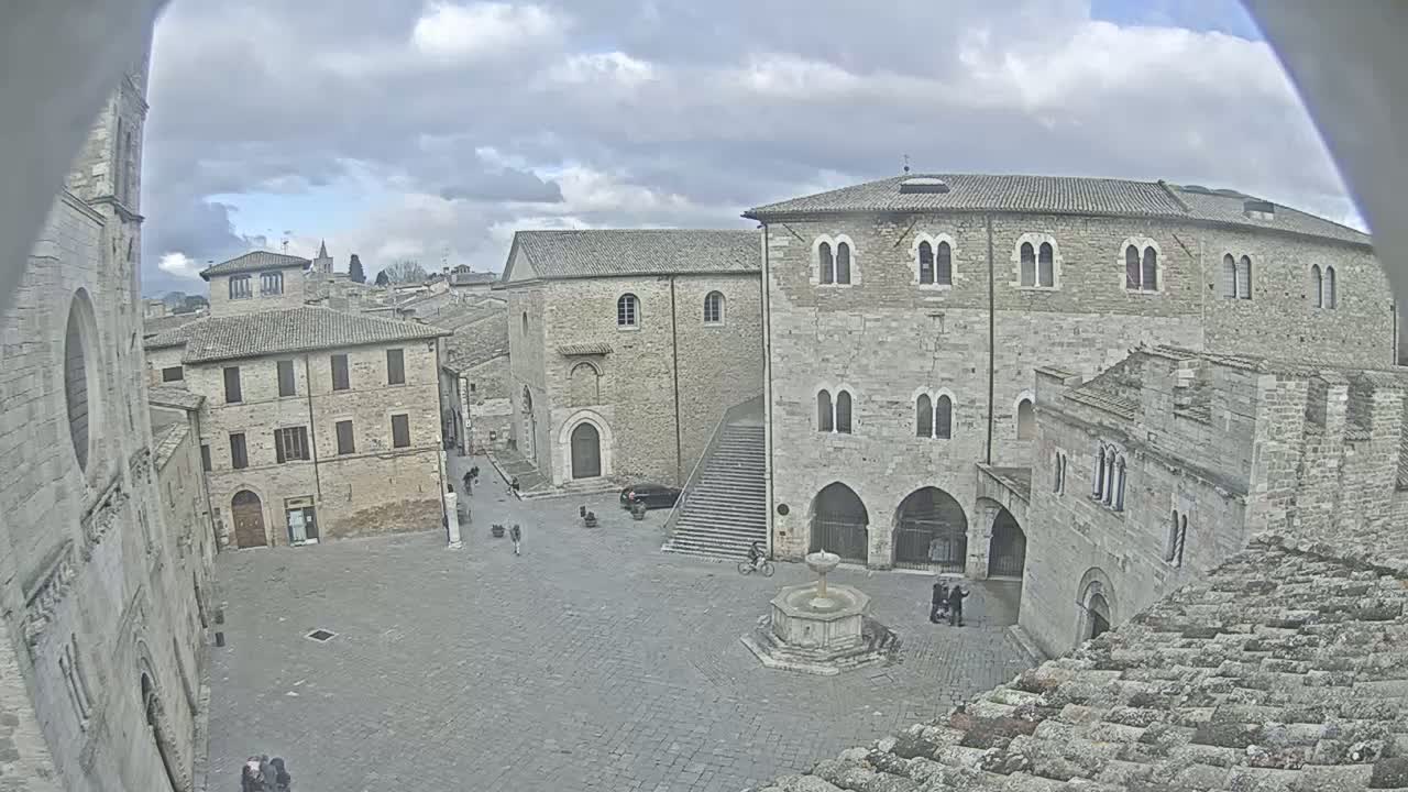 A stone plaza with a fountain is surrounded by medieval buildings under a cloudy sky.